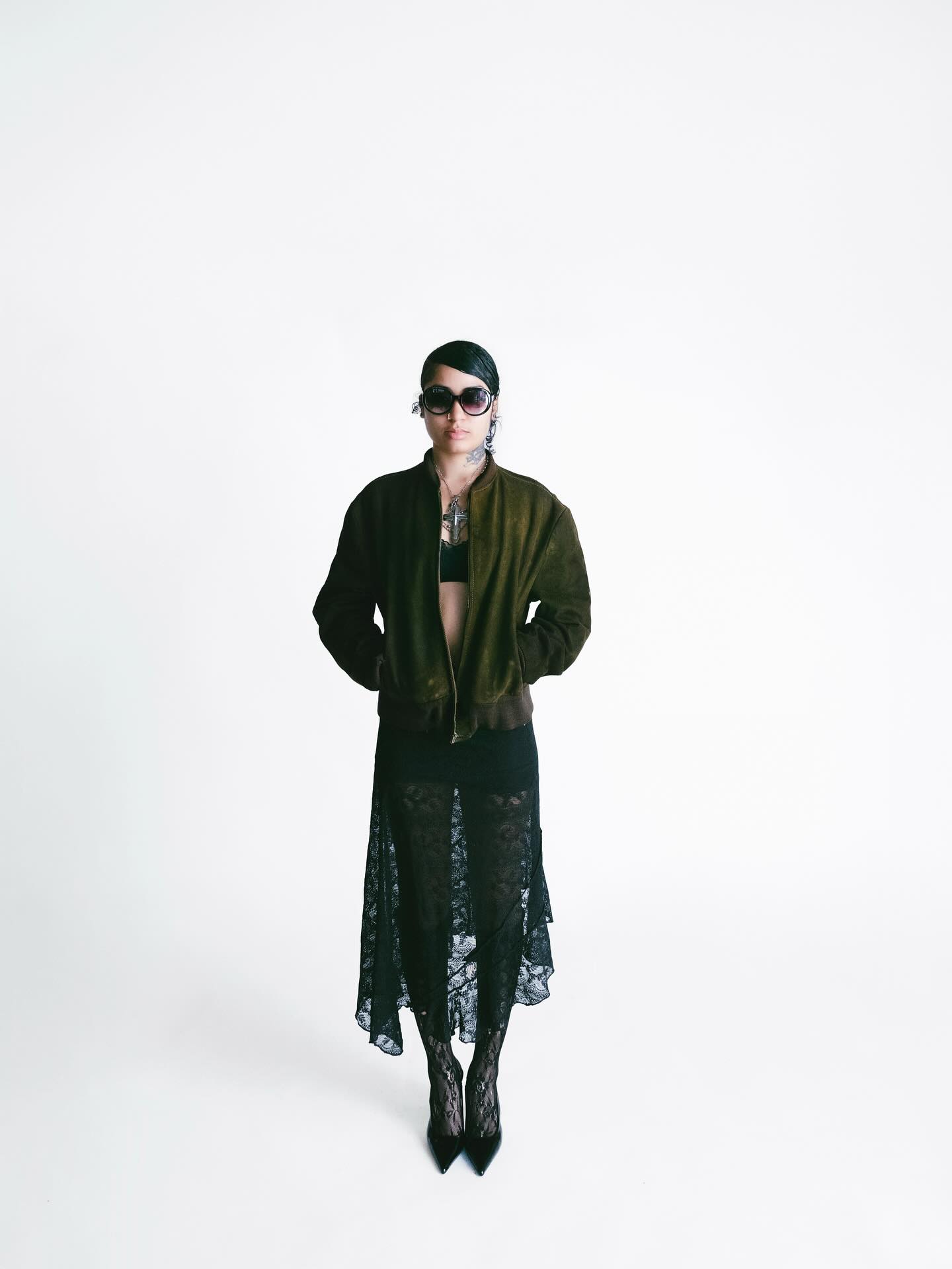 Lailah in sunglasses, an olive jacket and a black lace skirt